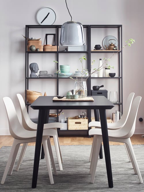 Zonă de sufragerie cu masă neagră, scaune, combinație de depozitare, lampă cu pandantiv, vază, plante, obiecte decorative, covor.
