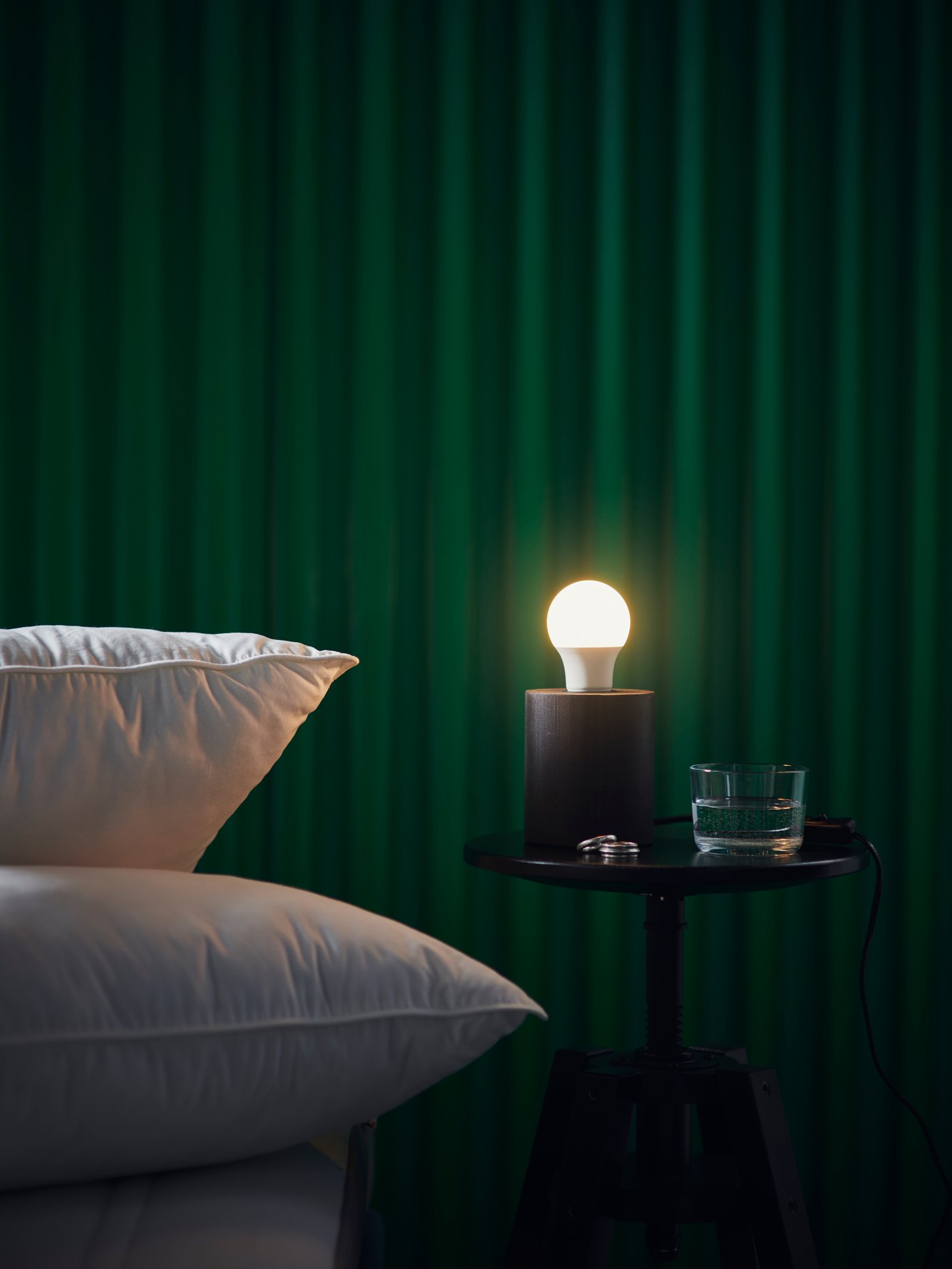 Twee witte kussens liggen naast een klein zwart tafeltje met een brandende SOLHETTA-lamp en een waterglas. De muur erachter is donkergroen. 