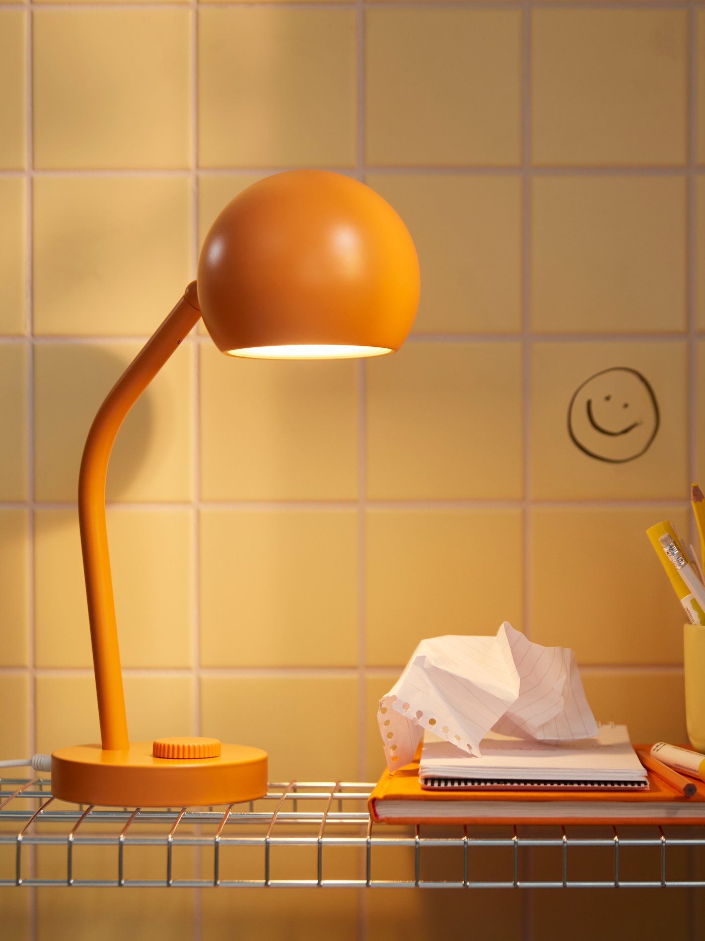 Lampada da scrivania LED KORVSNÖRE arancione dimmerabile accesa su un ripiano metallico, accanto a quaderni e portapenne colorato.