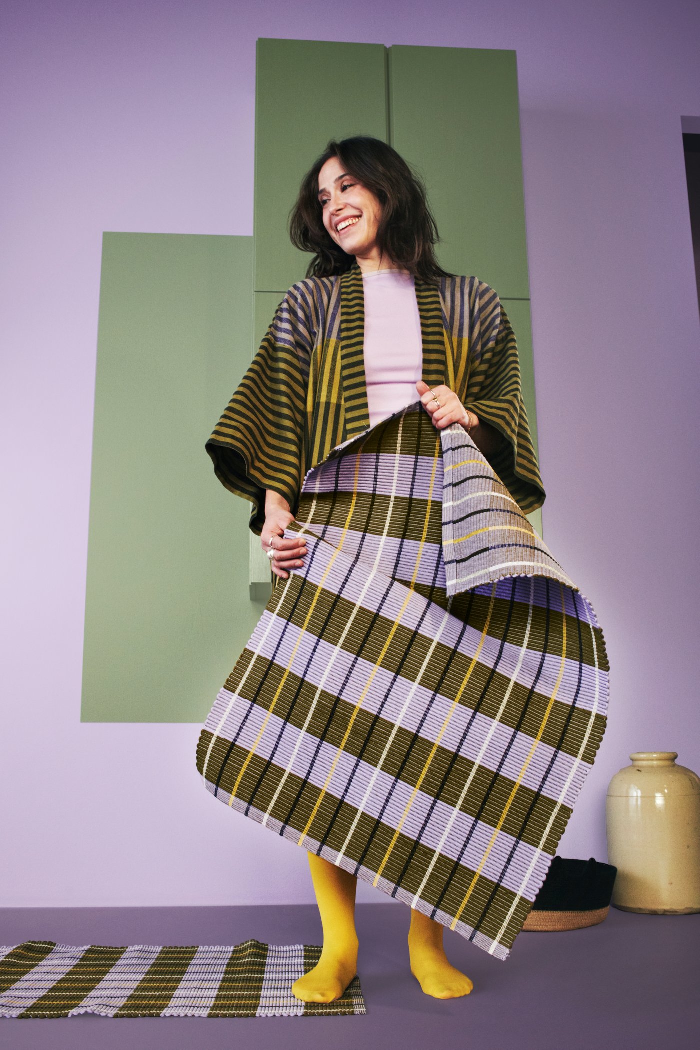 Una persona con una bata MÄVINN sujetando una alfombra de rayas MÄVINN de varios colores.