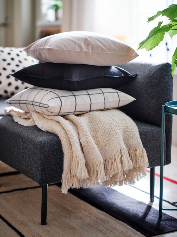 A cushion with a SPIKKLUBBA cushion cover and two other cushions are piled on top of a HUMLEMOTT throw on a sofa.