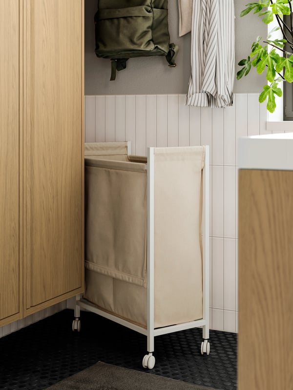 A white ENHET laundry bag with castors is placed in a corner next to an open HAGAÅN wall cabinet with oak effect.
