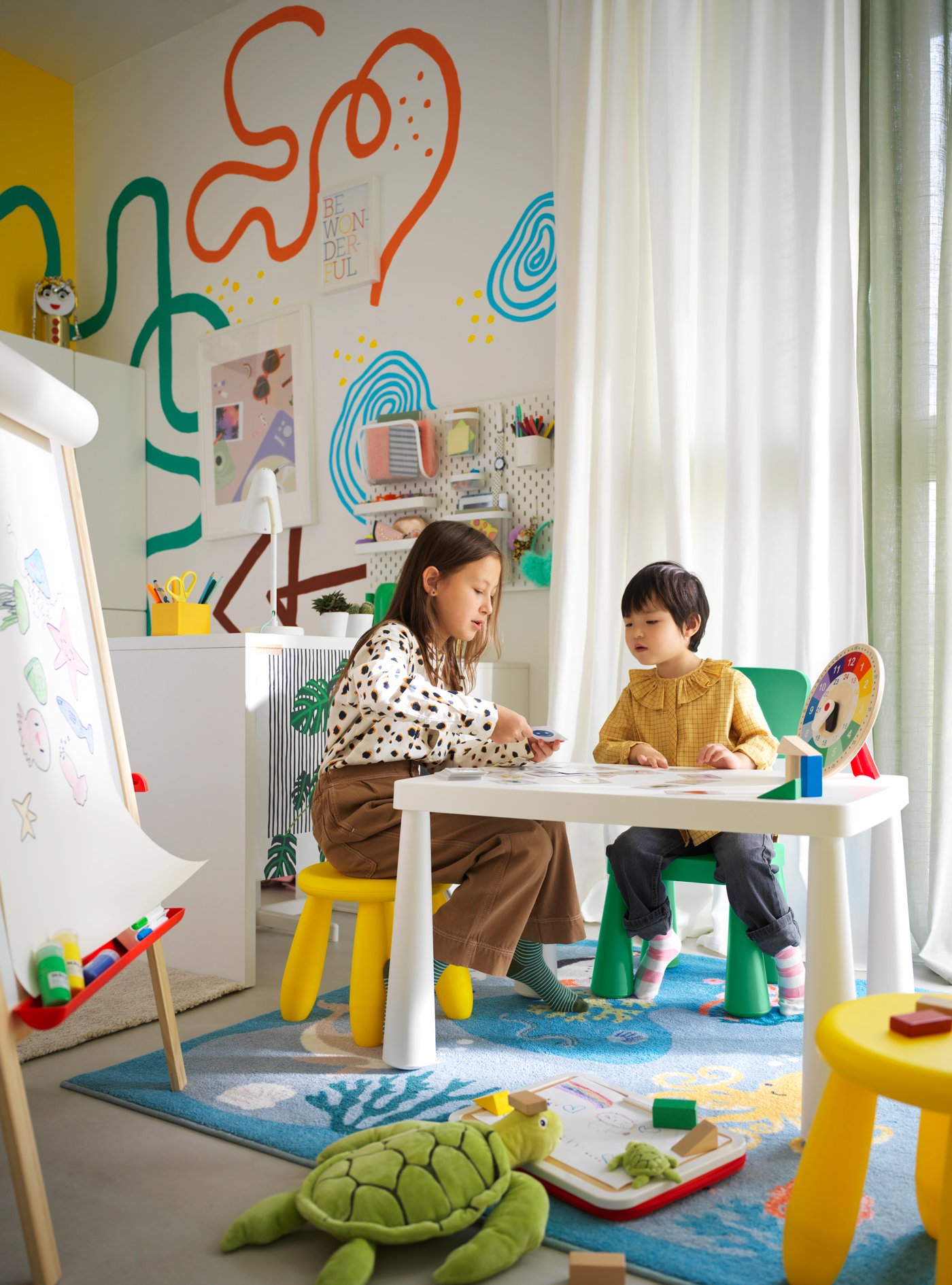 A child sits on a green MAMMUT children’s chair at a table with an adult who is sitting on a yellow MAMMUT children’s stool.