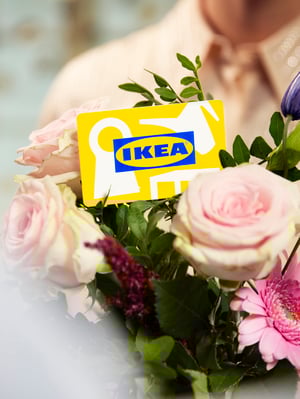 Buchet de flori cu trandafiri roz și alte flori colorate, ținut în mâini. În mijlocul buchetului se află un card galben cu logo-ul IKEA.