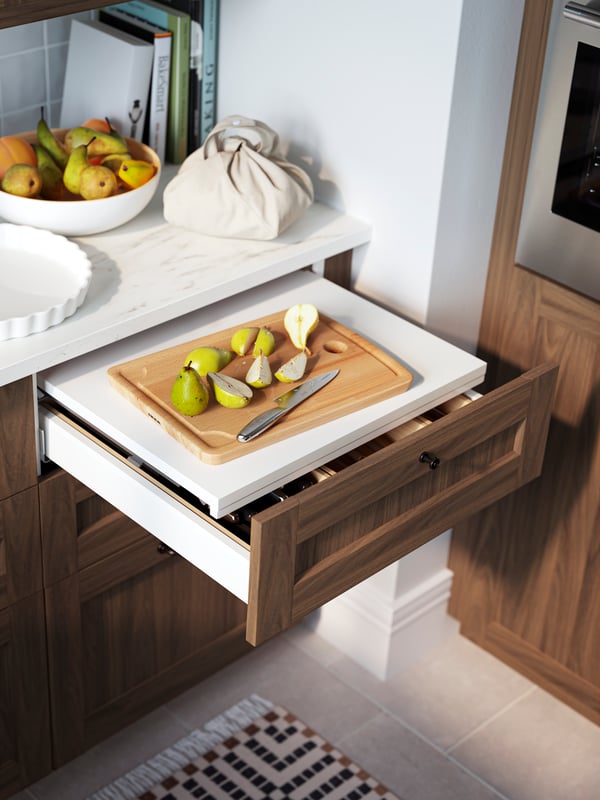 An open kitchen drawer with a brown walnut-effect ENKÖPING front with an integrated shelf inside used as extra worktop space to place a chopping board.