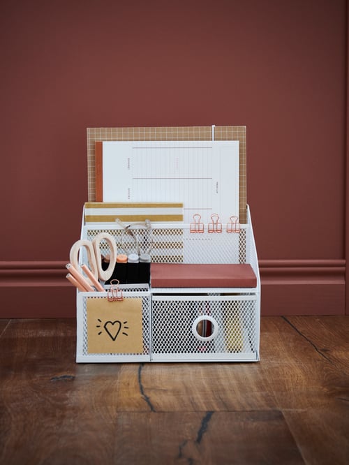 A desk organizer in white mesh with different compartments holding desk accessories like scissors, pens, papers and more.