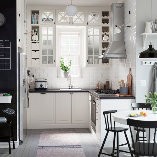 A kitchen with off-white glass-doored cabinets arranged around a window and a black mineral effect worktop.
