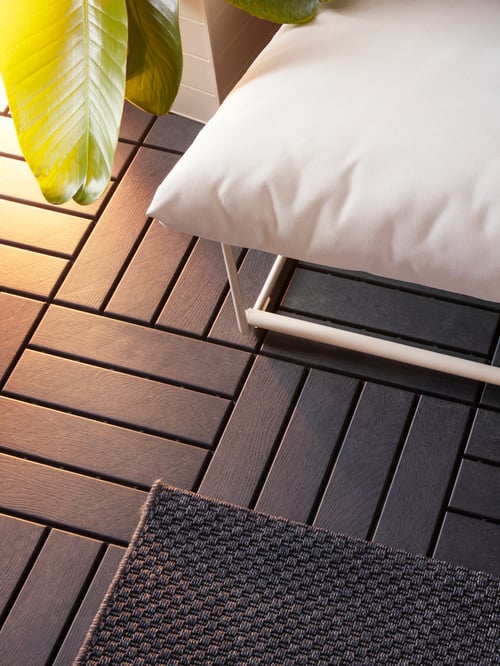 Pieces of dark grey RUNNEN outdoor floor decking with wide slats are under a chair and a rug in an outdoor area.