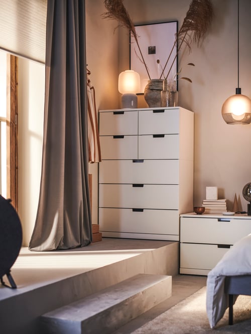 Bedroom area featuring a white chest of drawers, a chest of two drawers, a pair of curtains, vases, and a lit lamp.