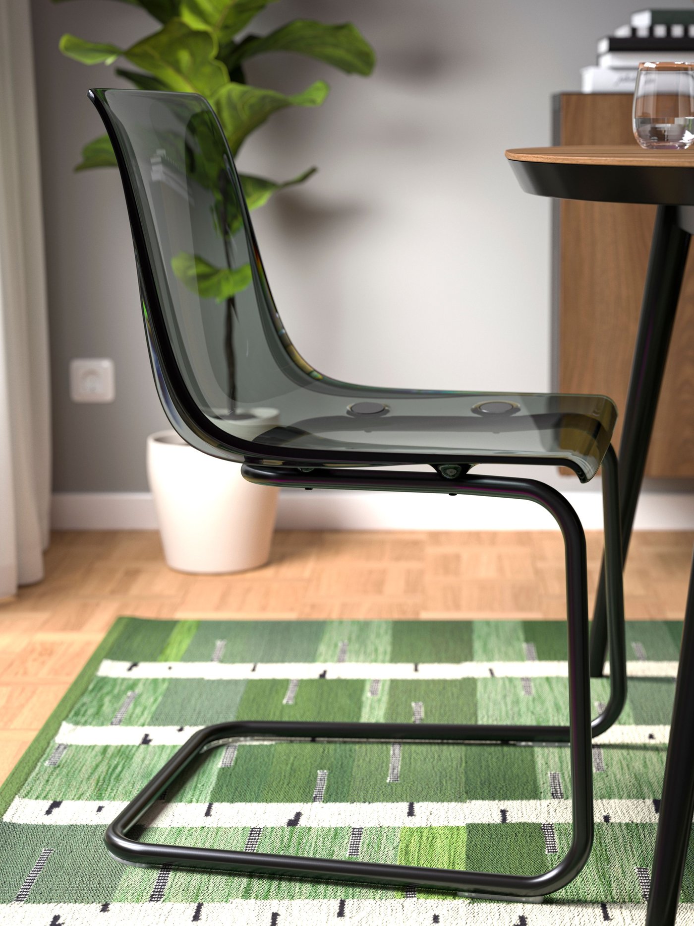 Une chaise transparente au profil moderne se détache devant un tapis vert à motifs géométriques, près d’une table aux pieds noirs et d’une plante en pot.