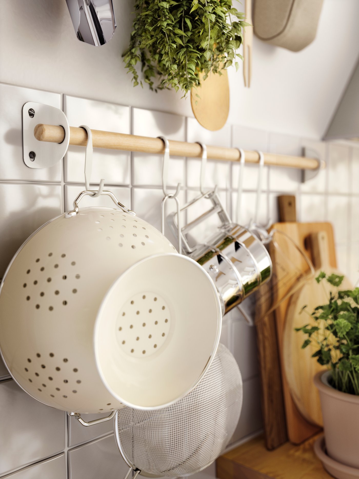 Köksredskap på krokstång, inklusive durkslag och sil, med örter och skärbrädor i bakgrunden.