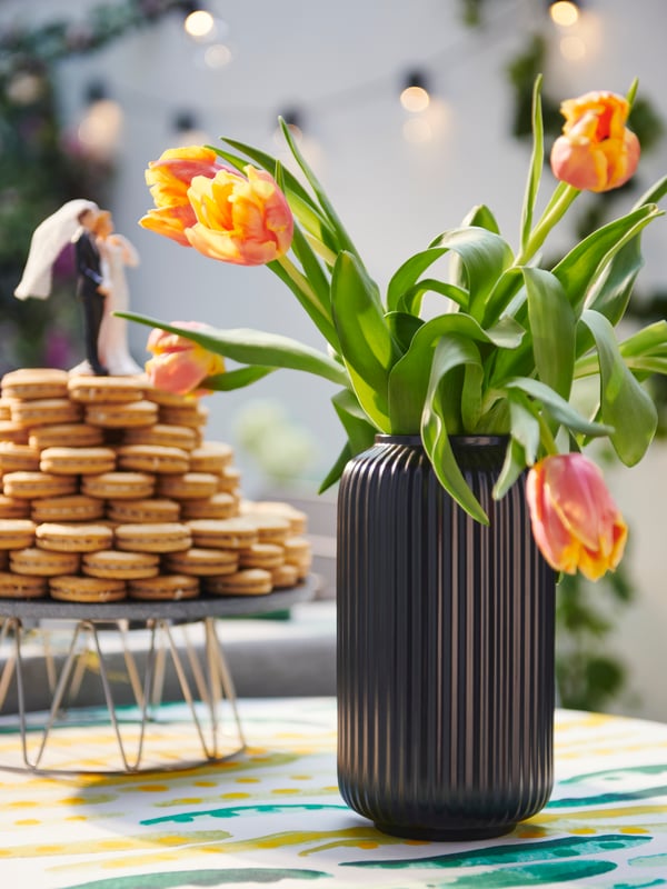 Sonnenbestrahlter Tisch mit einem BAKGLAD Kuchenteller, auf dem KAFFEREP Kekse zu einer Hochzeitstorte gestapelt sind. Daneben steht eine STILREN Vase mit Tulpen.