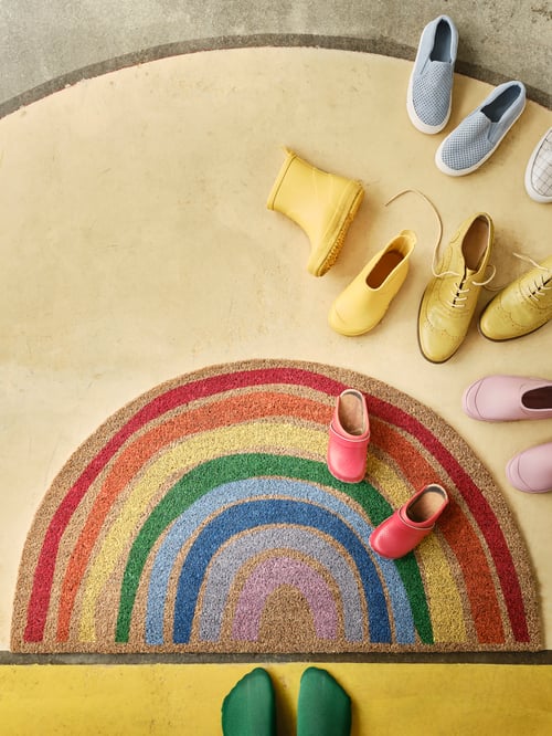 PILLEMARK door mat with a rainbow pattern lying on a yellow floor with colourful shoes and a pair of feet with green socks.