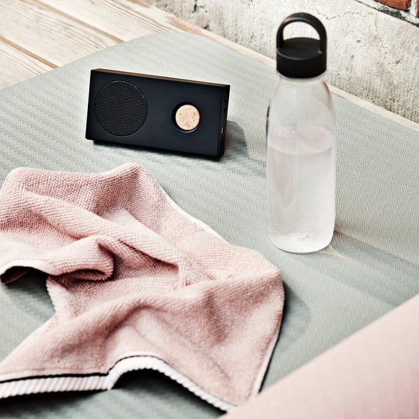 An IKEA 365+ water bottle with a black cap on a grey gym mat, a pink towel over it and a small Bluetooth speaker in black.