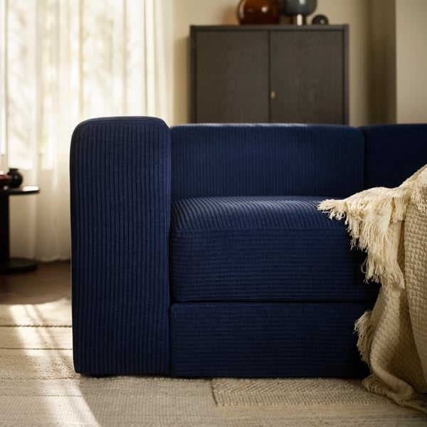 A LAPPTÅG throw on a dark-blue JÄTTEBO four-seat sofa in a living room with sunlight sifting through STENFRÖ sheer curtains.