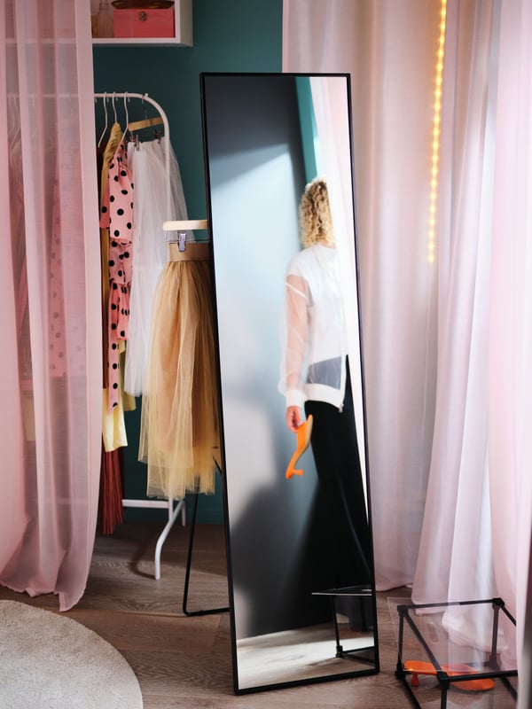 A person reflected in a black KARMSUND standing mirror that’s in front of a pink STENFRÖ curtain and a white clothes rail.