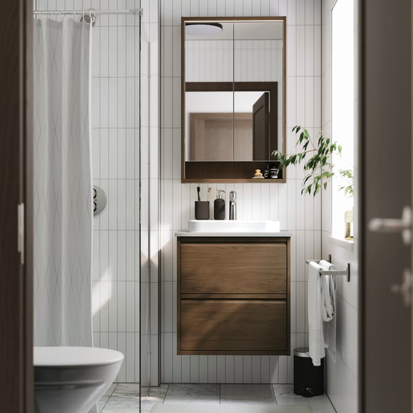 An ÄNGSJÖN vanity sink in brown oak effect is placed in a white bathroom with a window and it has a mirror cabinet above it.