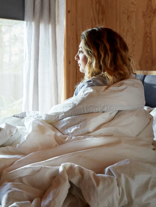 Bedroom featuring a person covered by a white duvet cover with 2 pillowcases made of cotton, looking outside.