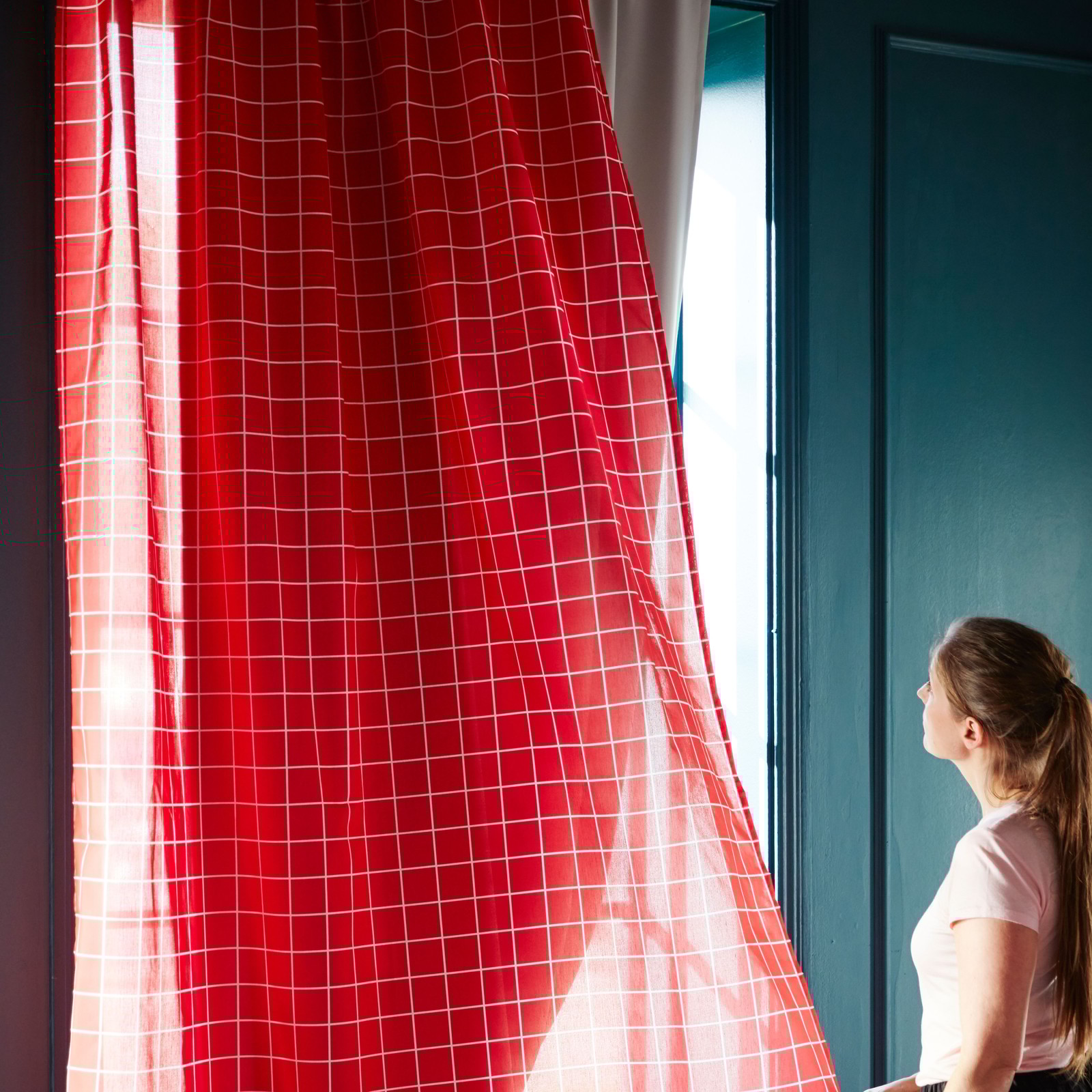 O femeie ține draperii roșii și privește afară, în timp ce lumina soarelui intră pe fereastră.