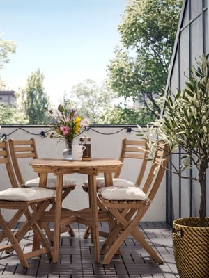 Măsuță rotundă și scaune pliante din lemn pe un balcon IKEA, cu perne albe, ghirlandă luminoasă și ghiveci cu măslin, pregătite pentru un mic dejun relaxat în aer liber.