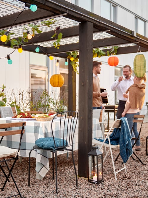 An outdoor scene with a group of people socialising together, a table set for a party, chairs and hanging lighting.