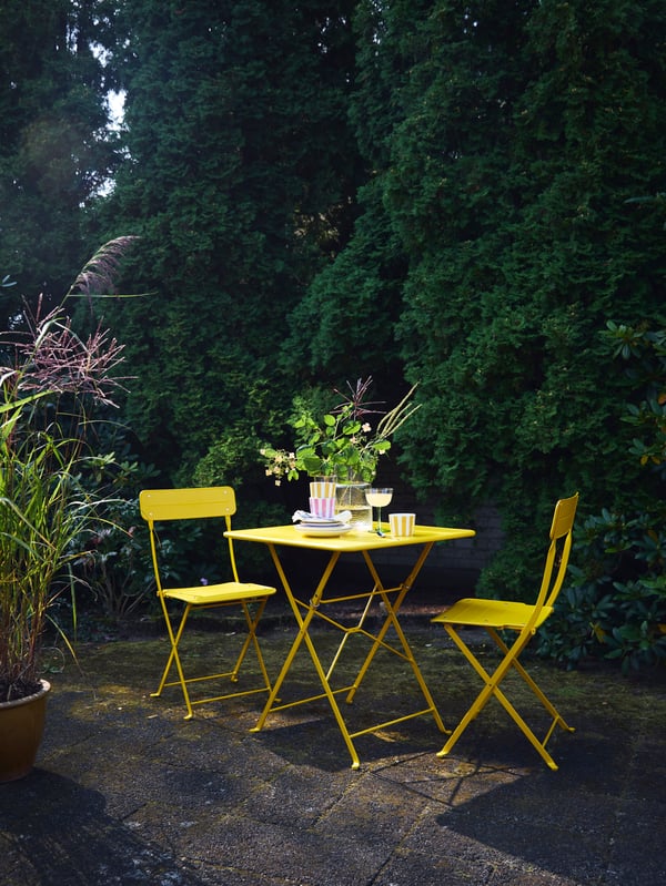 SUNDSÖ outdoor table for two in light yellow brightens a backyard in an evening setting.