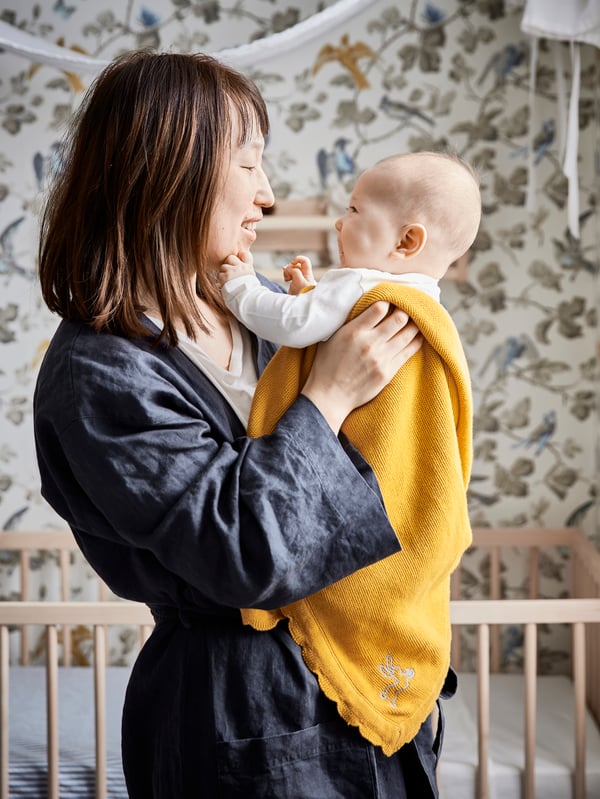 Une mère debout dans un peignoir tenant son bébé enveloppé dans une couverture jaune.