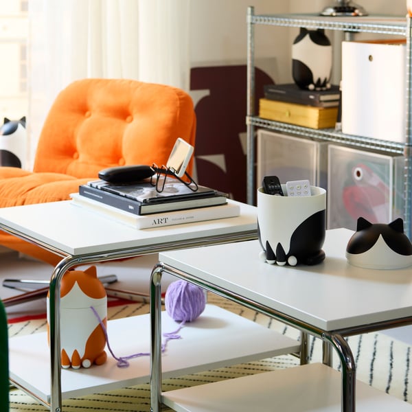 Three cat-shaped GRESIMOJS storage with lid are on tables and shelves in a room. Two are black/white. One is orange/white.