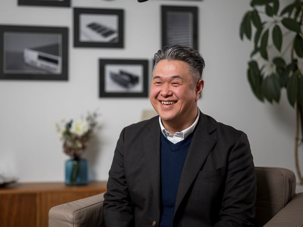 ジャケットを着た人物が、アートと観葉植物を背景にした落ち着いたオフィスの一角に座っている。