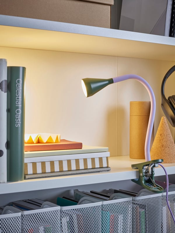 A lilac/dark green NÄVLINGE LED clamp spotlight is attached to a shelf and shines its light on some books.