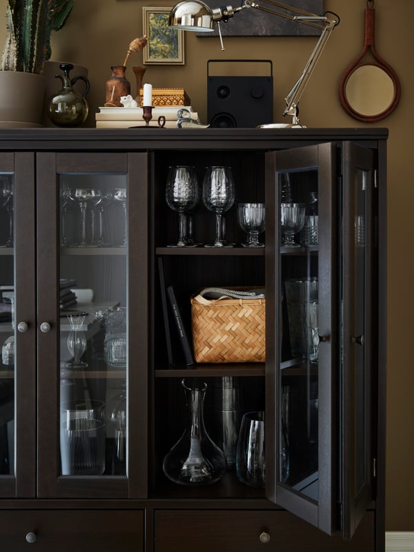 An IDANÄS cabinet with bi-folded glass doors containing glassware and a basket stands against a wall with one door open.