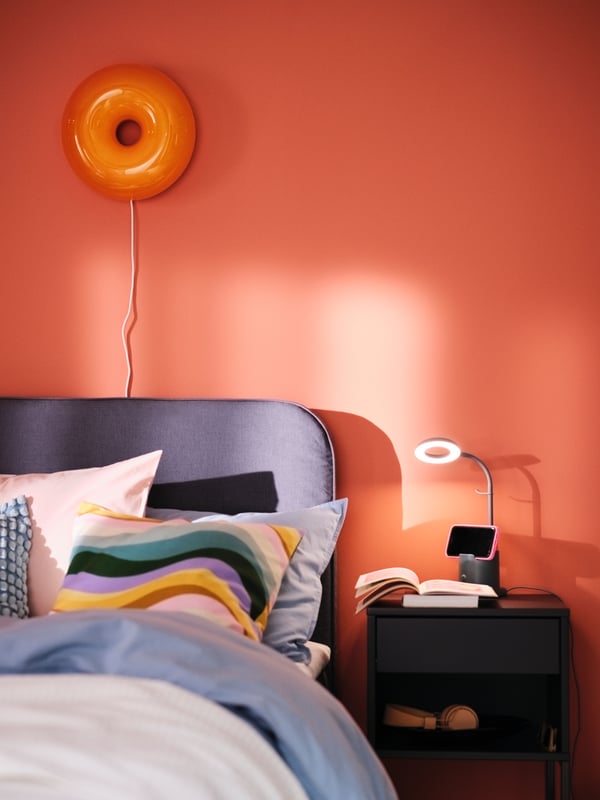 A BLÅFJÄLLET ottoman divan bed with a VARMBLIXT LED table/wall lamp on the wall behind it and colourful cushions on top.