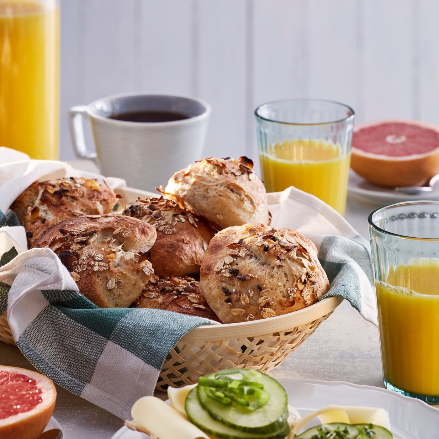 Un panier de petits pains confectionnés avec du muesli HJÄLTEROLL sur une table de petit déjeuner, près d’une tasse de café, d’un jus d’orange et d’un demi-pamplemousse.