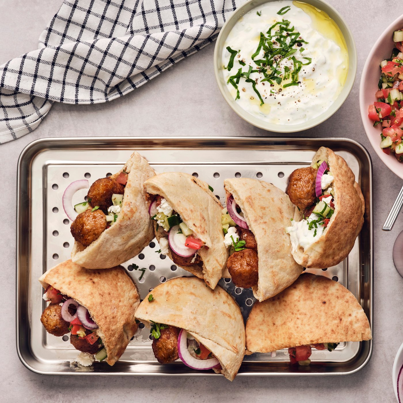 Eight pieces of pita bread are filled with meatballs and tzatziki, placed on a metal tray next to a towel and two bowls.