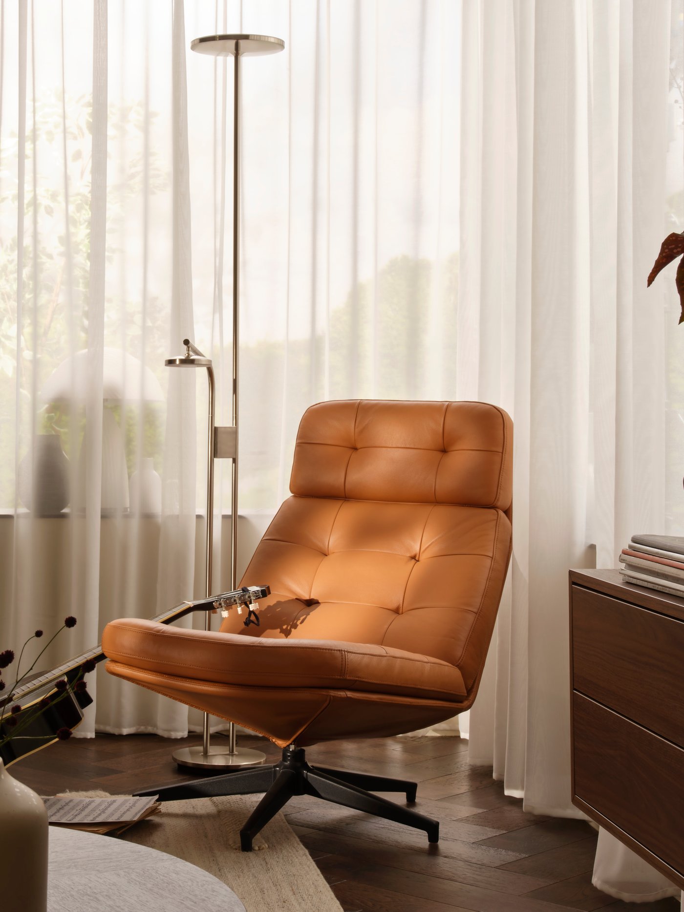 Ett hörn av ett vardagsrum med en gyllenbrun HAVBERG snurrfåtölj bredvid ett stort fönster med STENFRÖ skira gardiner.