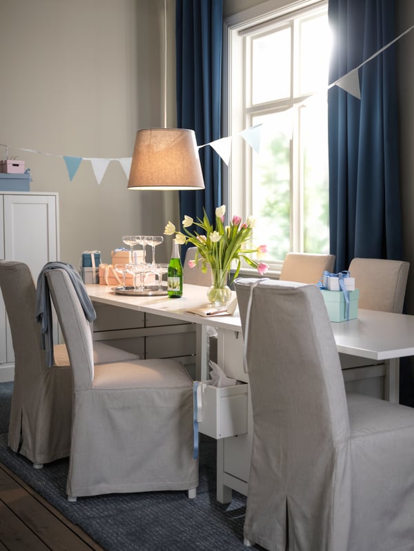 Una zona de comedor decorada con una mesa blanca NORDEN y sillas BERGMUND con fundas largas de color beige y gris oscuro.
