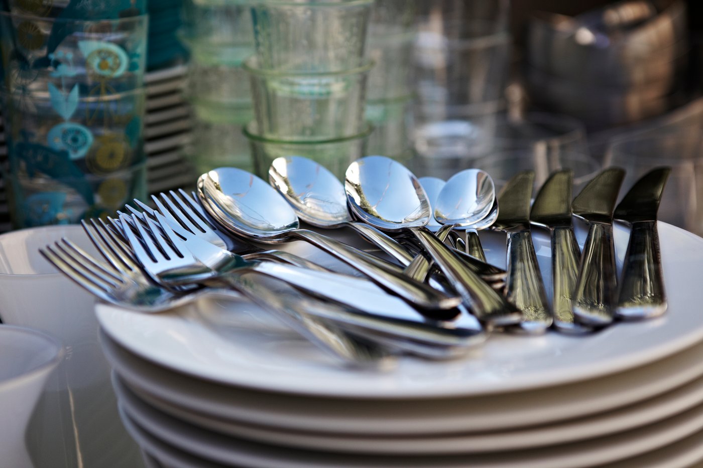 Ein Stapel weisser Teller mit glänzendem Besteck aus Löffeln, Gabeln und Messern, umgeben von Gläsern und weiterem Geschirr im Hintergrund.