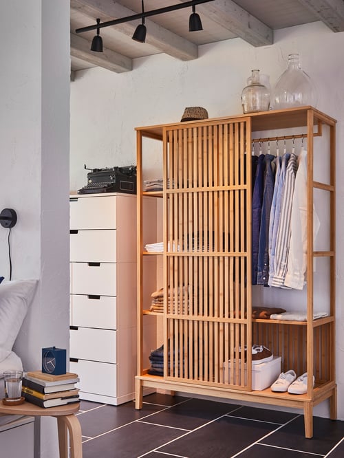 By a chest of drawers is a bamboo NORDKISA open wardrobe with its one sliding door revealing clothes on shelves and a rail.
