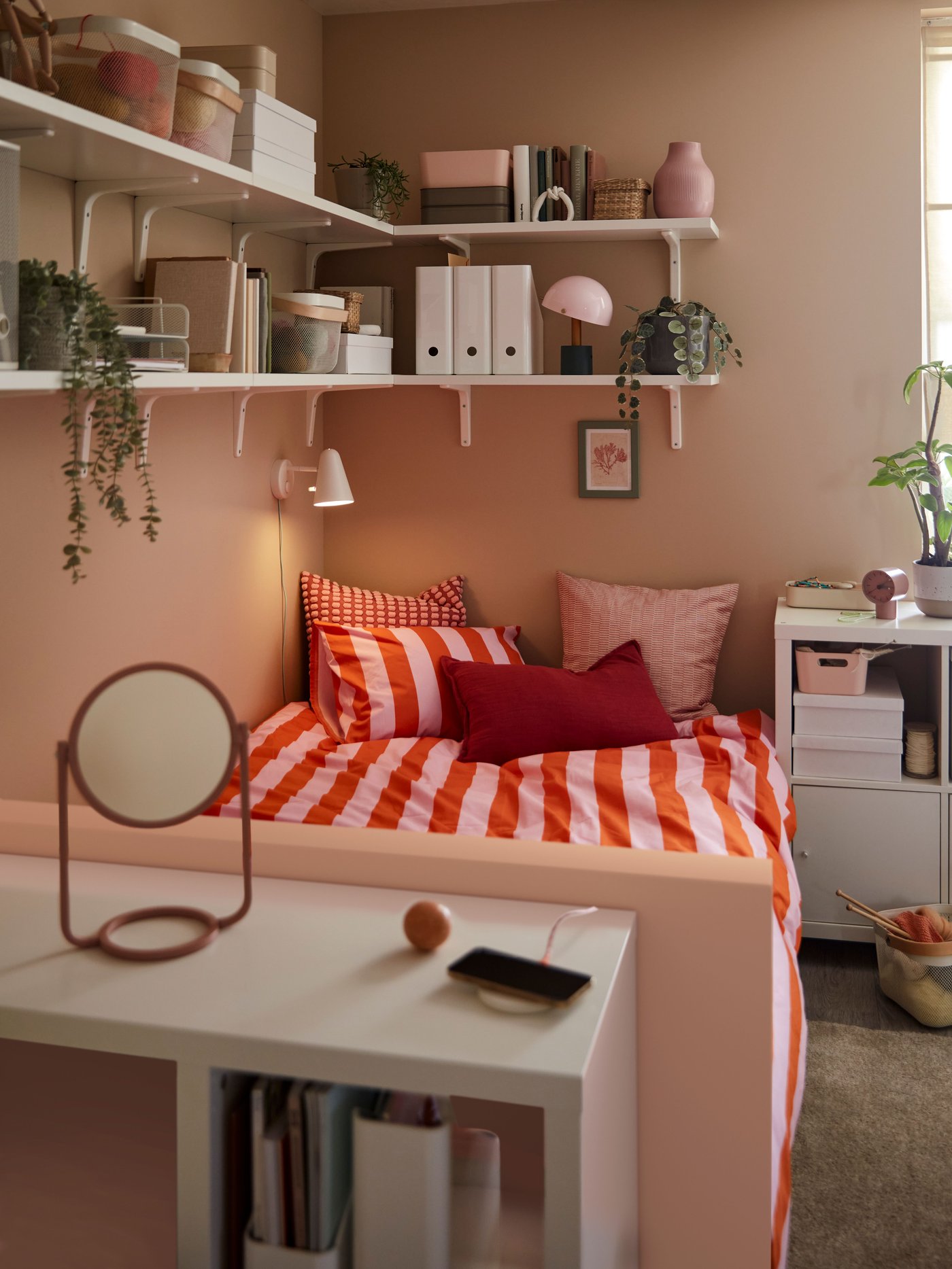 Ett litet rum med ett vitt KALLAX skrivbord vid fotändan av en säng med färgglada sängkläder och vägghyllor ovanför.