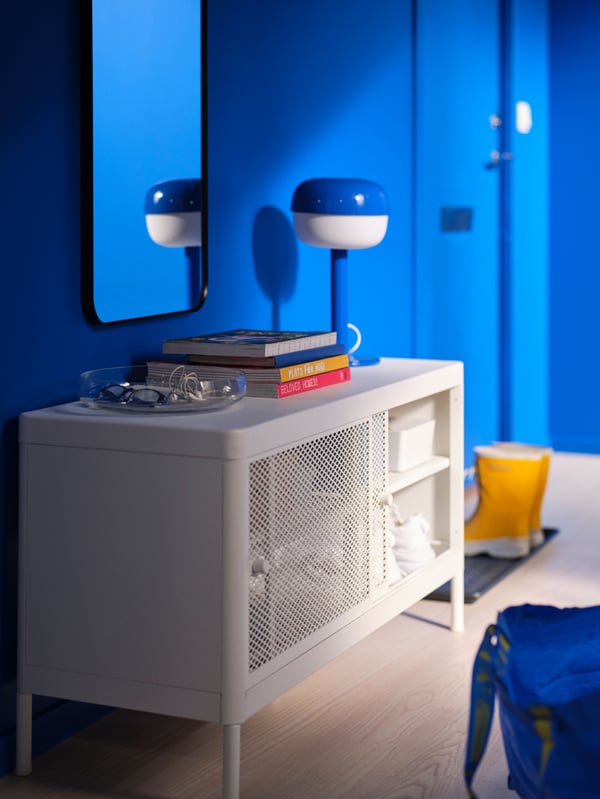 A white MACKAPÄR shoe cabinet in a blue hallway with a blue and white BLÅSVERK table lamp on top and a pile of books.