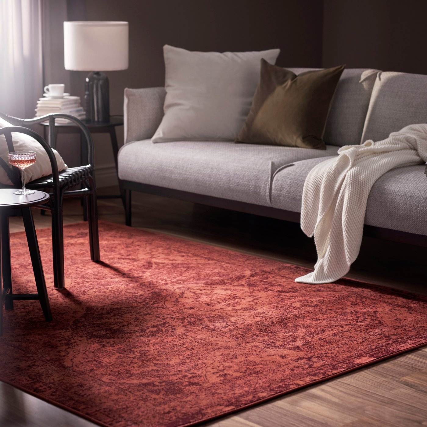 Vardagsrum med grå soffa, vita och bruna kuddar, röd matta, litet bord med vin och böcker, lampa i bakgrunden. 