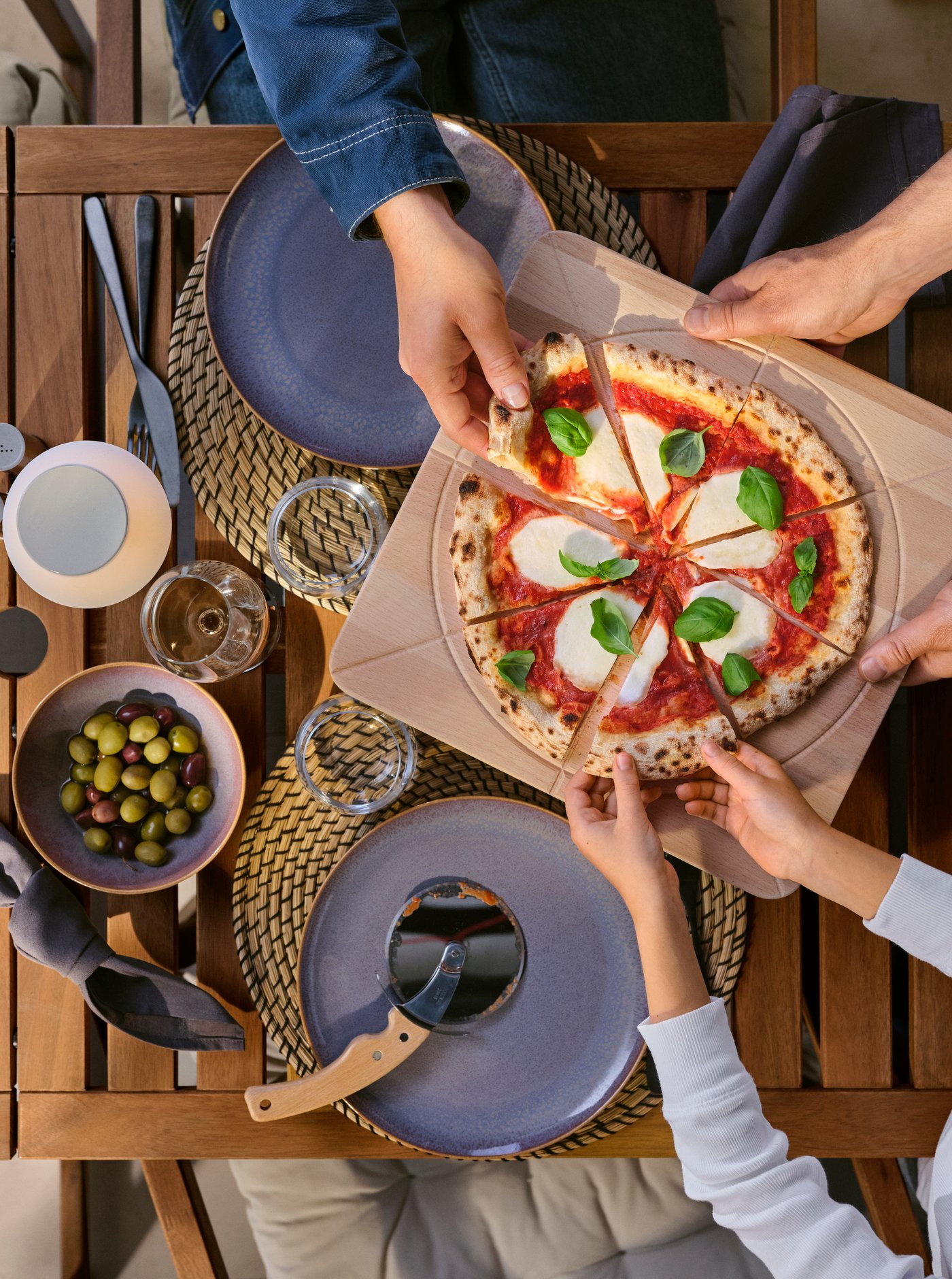 Ruce tří lidí si dělí pizzu na prkénku SÅPÖRTMAL u venkovního stolu, podávanou na nádobí FÄRGKLAR.