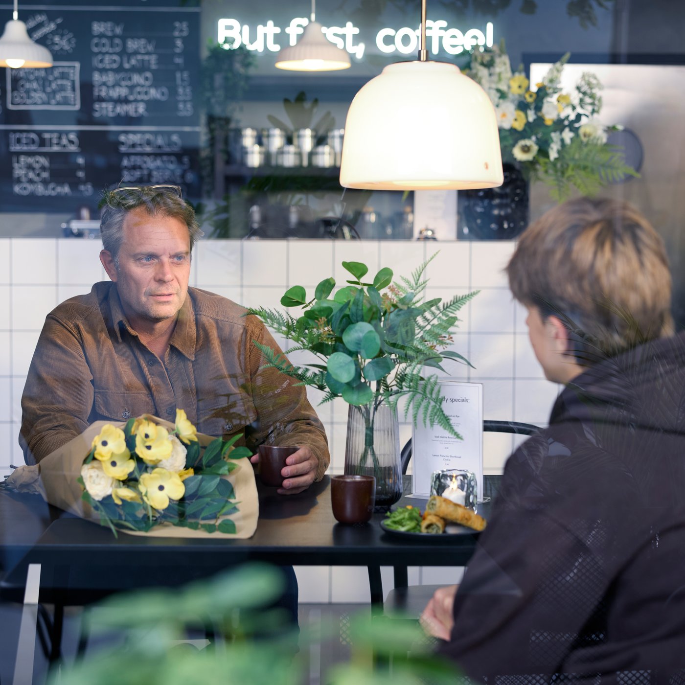 Zicht door een raam van een café met twee mensen die aan een LISABO tafel in zwart essen koffie drinken. Rond hen zien we planten en bloemen.