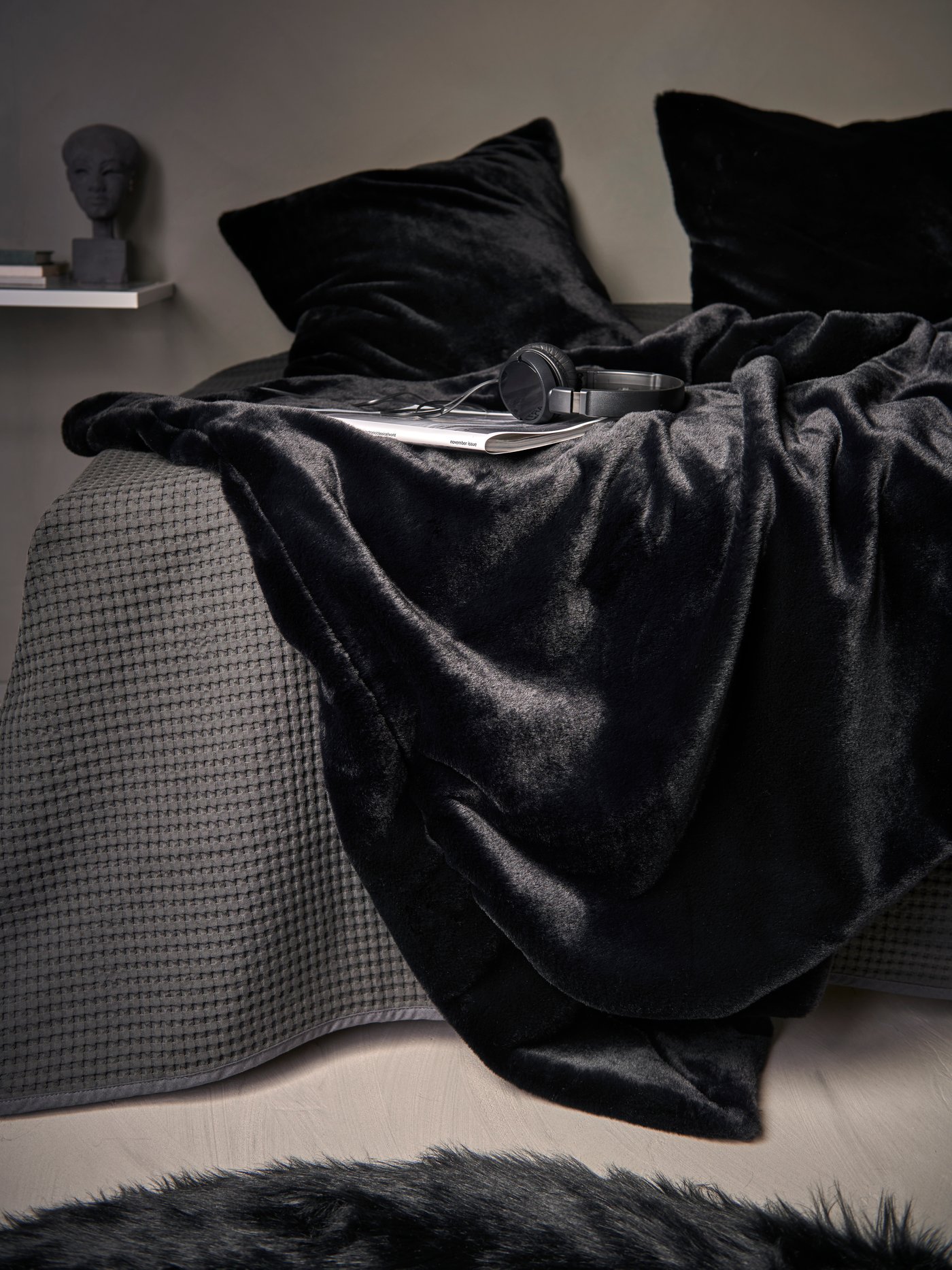 A black OBEGRÄNSAD throw and cushion covers draped on a grey waffle bedspread with headphones and magazine lying on top.