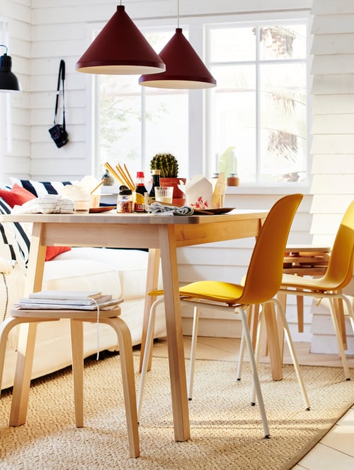 Salle à manger avec table rectangulaire en bouleau placée sous 2 suspensions, à côté de 2 chaises jaunes et d’un tabouret.