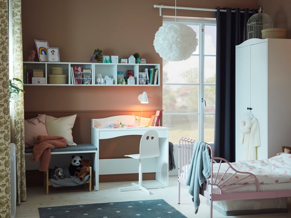A children's bedroom with a light pink extendable bed frame with slatted bed base, white desk, books and soft toys.