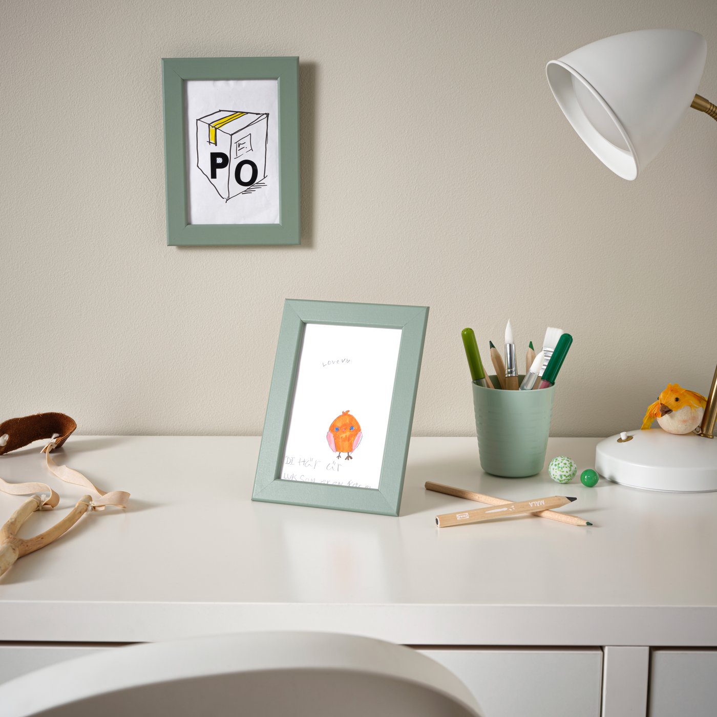 Cornice FISKBO verde grigio chiaro appoggiata su una scrivania bianca, con illustrazione colorata all’interno.