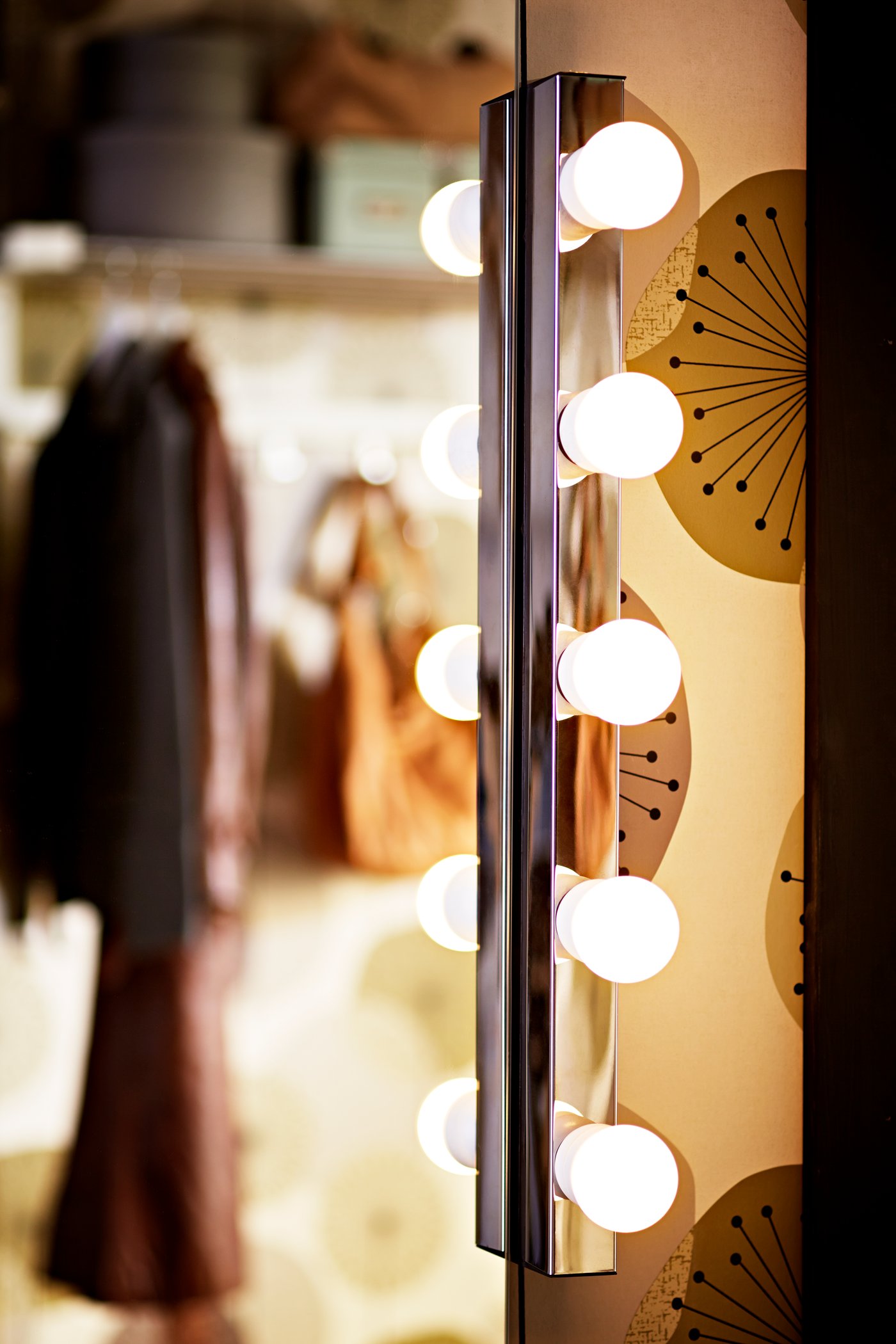 En vertikal MUSIK lampa med runda glödlampor lyser mot en spegel i ett rum med mönstrad vägg och klädförvaring i bakgrunden.