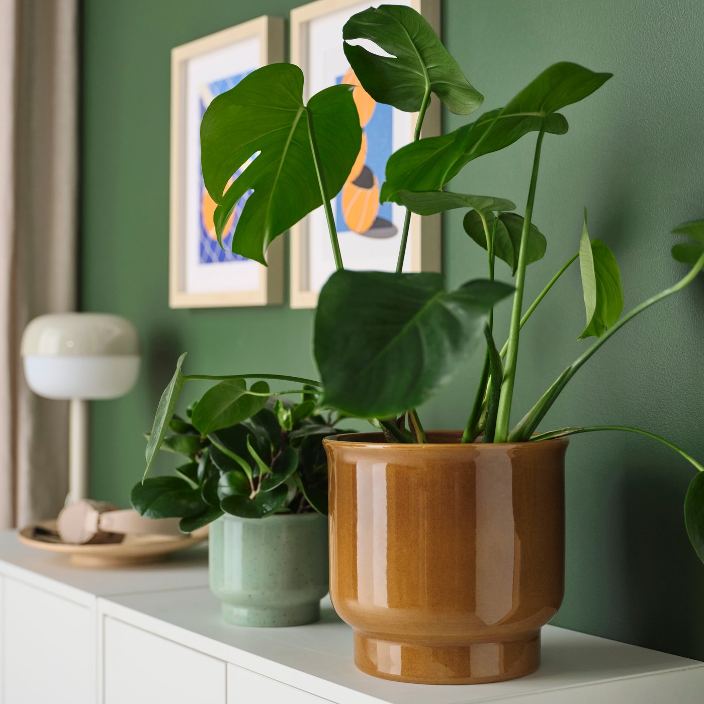 A MONSTERA plant in a yellow‑brown SAMMETSSYREN plant pot next to a blue flower pot on a sideboard in the living room.
