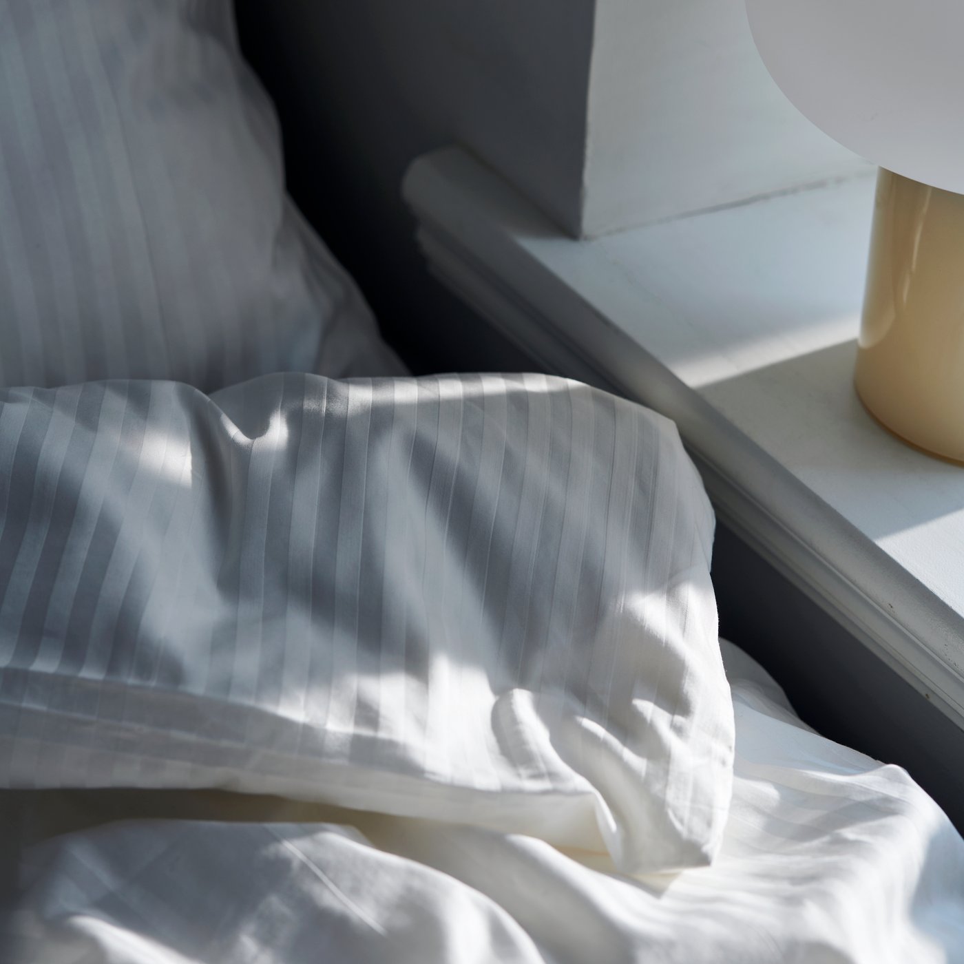 The corner of a folded-over duvet covered in NATTJASMIN bed linen in white in a sunny space by a shelf.